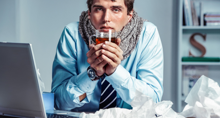 Das Bild zeigt einen Mann in Businesskleidung, der krank aussieht und mit müdem Blick eine Tasse Tee in den Händen hält. Er trägt einen dicken Schal um den Hals, während er an einem Schreibtisch sitzt, umgeben von zerknüllten Taschentüchern und einem Laptop.
