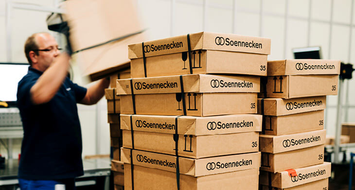 Im Vordergrund sieht man mehrere Pakete der Firma Sonnecken aufeinandergestapelt. Im Hintergrund sieht man verschwommen einen Mitarbeiter, der einen weiteren Karton auf den Stapel hebt.