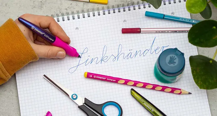 Auf einem Collegeblick steht das Wort „Linkshänder“. Eine Hand hält mit links einen Linkshänder-Schreiblernfüller, drumherum liegen weitere Produkte für Linkshänder.