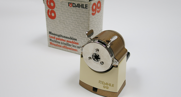 Das Bild zeigt einen klassischen, mechanischen Bleistiftspitzer von Dahle, Modell 99, zusammen mit seiner Originalverpackung. Der Spitzer hat eine cremefarbene Basis und eine Metallfront mit zwei Hebeln.“