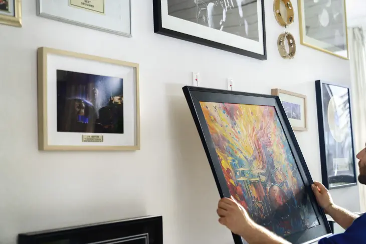 Ein Bild von einem Mann, der mit tesa-Klebenägeln ein Bild an die Wand hängt. An der Wand hängen bereits andere Bilder in verschiedenen Formaten. 
