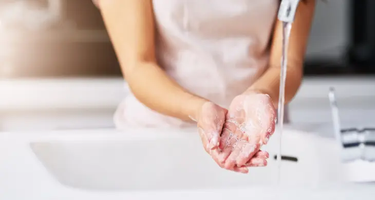 Das Bild zeigt den Oberkörper einer weiblichen Person, die sich die Hände unter einem Wasserhahn wäscht. Die Hände sind mit Seife bedeckt, während Wasser darüber fließt. Im Hintergrund ist ein Teil des Waschbeckens und des Wasserhahns zu sehen.