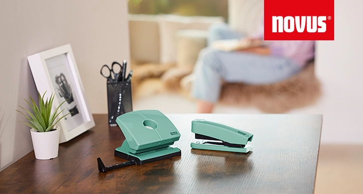 Auf einem Holzschreibtisch stehen ein grüner Locher und ein grünes Heftgerät von Novus sowie eine kleine Topfpflanze, ein Stiftehalter mit Scheren und ein eingerahmtes Schwarz-Weiß-Foto. Im Hintergrund ist eine Person auf einem Sofa zu sehen.
