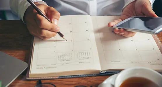 Eine Hand schreibt in einen analogen Kalender, die andere Hand hält ein Smartphone fest. 