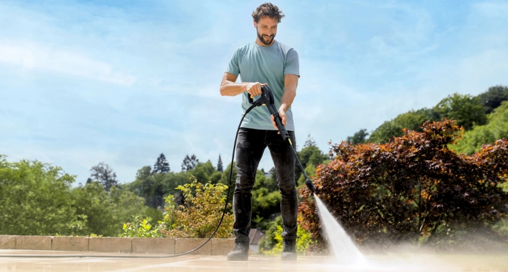 Mann reinigt eine Terrasse mit einem Kärcher Hochdruckreiniger, umgeben von Sträuchern und Bäumen.