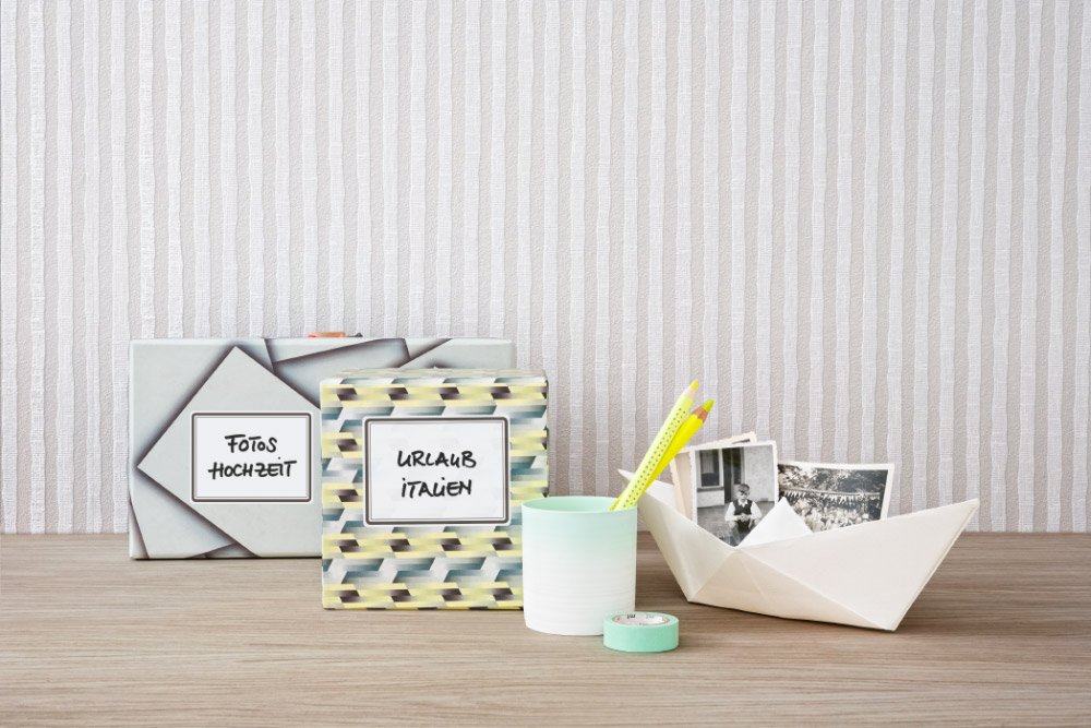 Auf einem Holztisch stehen 2 Aufbewahrungsboxen für Fotos, welche mit Avery Zweckform Living Etiketten beklebt und mit der Aufschrift "Fotos Hochzeit" und "Urlaub Italien" beschriftet wurden. Daneben stehen ein Stifteköcher und ein aus Papier gefaltetes Schiff, in dem Fotos liegen.