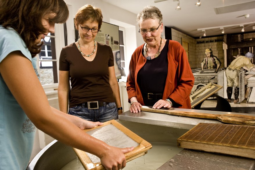 Zwei Frauen schauen im Papiermuseum dabei zu, wie Papier geschröpft wird. 