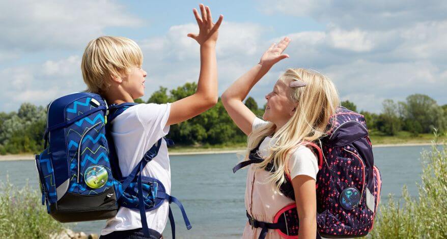 Ein Junge und ein Mädchen mit ergobag Schulranzen auf dem Rücken stehen vor einem See und geben sich High-Five.