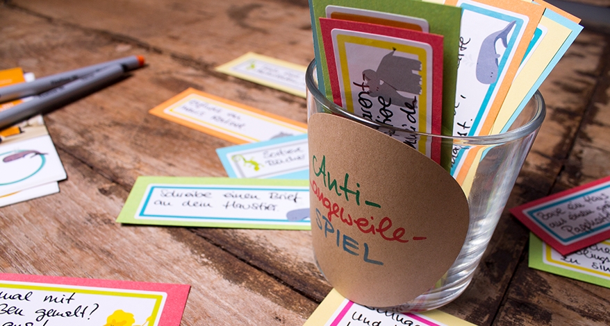 Nahaufnahme des Glases, welches mit bunten Aufgaben-Kärtchen bestückt ist. Weitere Aufgaben-Kärtchen liegen auf dem Tisch verteilt.