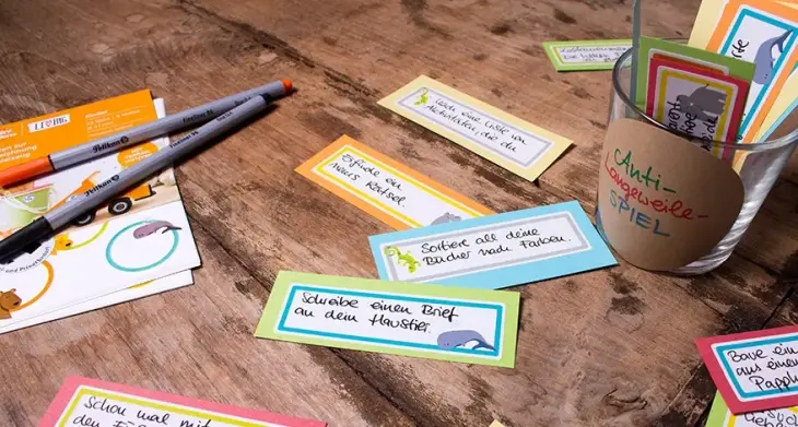 Auf einem dunklen Holztisch liegen selbstgebastelte bunte Kärtchen, welche mit Avery Zweckform Living-Etiketten zur Kennzeichnung von Spielzeug beklebt und mit kleinen Aufgaben beschriftet sind. Daneben steht ein Glas, das mit einem Aufkleber und der Aufschrift "Anti-Langeweile-Spiel" beschriftet ist. In dem Glas sind weitere bunte Kärtchen. Außerdem liegen auf dem Tisch 2 Fineliner und eine Stickerkarte mit den Aufklebern von Avery Zweckform.