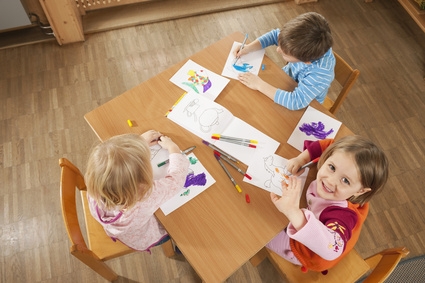 3 Kindergarten Kinder sitzen an einem Tisch und malen mit Buntstiften. Ein Kind schaut in die Kamera und lächelt.
