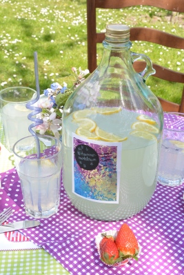 Auf einem mit lila und grün ausgelegtem Tisch in einem Garten steht eine große Glasflasche, welche mit Zitronenlimonade gefüllt ist. Auf der Flasche ist ein selbst gestaltetes Etikett aufgeklebt. Neben der Flasche stehen Gläser mit gekringelten Glas-Stohalmen.