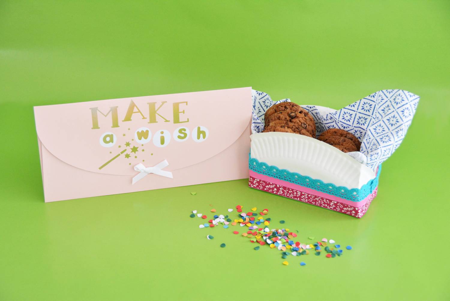 Eine selbstgebastelte Keksverpackung steht auf einem grünen Untergrund. Daneben steht ein rosa Umschlag mit der Aufschrift "Make a wish".