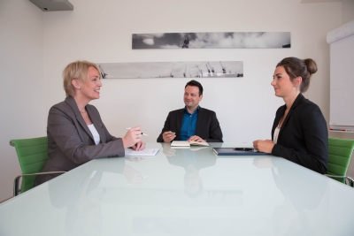 2 Frauen und ein Mann sitzen in einem Besprechungsraum und führen ein berufliches Gespräch. Alle haben Notitzbücher vor sich liegen und einen Füller in der Hand.