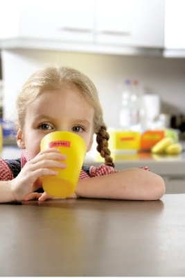 Ein kleines Mädchen sitzt an einem Tisch und trinkt aus einem gleben Becher, der mit einem roten Etikett von Dymo beschriftet ist.
