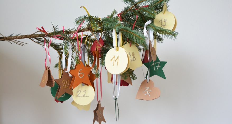 An einem Tannenzweig hängen runde und sternförmige Adventskalender Anhänger aus Papier. 