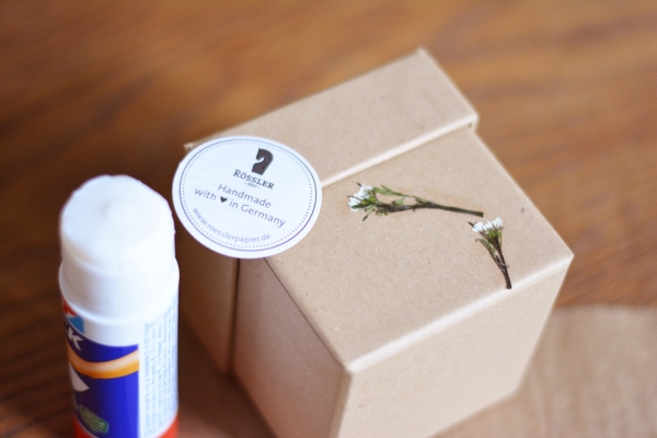 Eine getrocknete Blüte wurde auf eine Box von Rössler geklebt, daneben steht ein Klebestift.