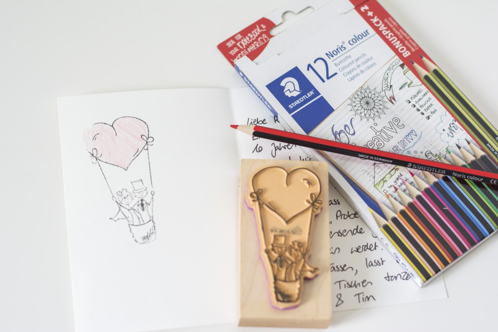 Auf einem weißen Tisch liegt eine geöffnete handgeschriebene Karte. Auf der linken Seite ist ein Stempel mit einem Brautpaar in einem Heißluftballon aufgedruckt. Der Ballon hat eine Herzform, welche mit einem roten Buntstift ausgemalt ist. Auf der rechten Seite der Karte liegt der Stempel und eine Packung Noris colour Buntstifte der Marke Staedtler, der rote Buntstift liegt auf der Verpackung.