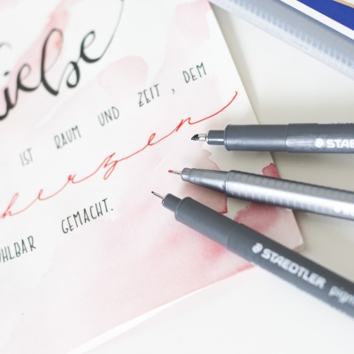 Nahaufnahme eines Ausschnitts einer handgeschriebenen Hochzeitskarte. Die Karte ist mit rter Aquarelltechnik bemalt, darauf ist mit schwarzem und rotem Fineliner der Marke Staedtler ein Spruch gelettert. Auf der Karte liegen 3 geöffnete Fineliner der Marke Staedtler.
