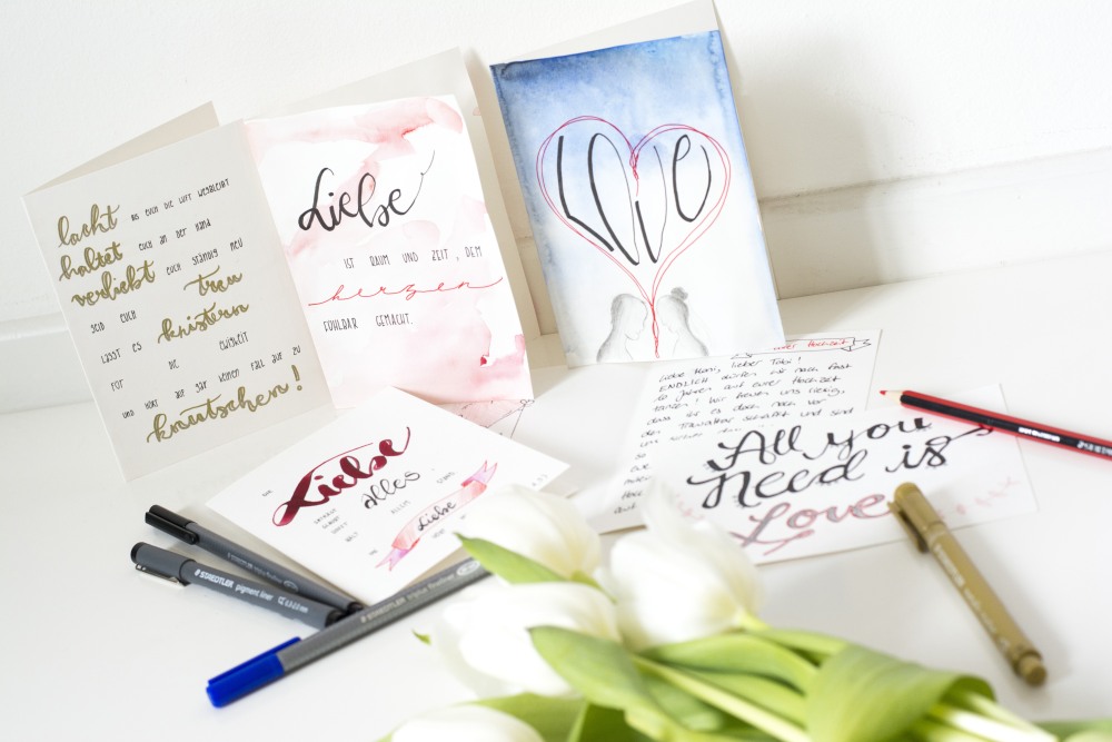 Auf einem weißen Tisch liegen und stehen diverse selbstgebastelte Karten, die mit Motiven und Sprüchen für eine Hochzeit gestaltet sind. Auf dem Tisch liegen mehrer Filzstifte und weiße Tulpen.