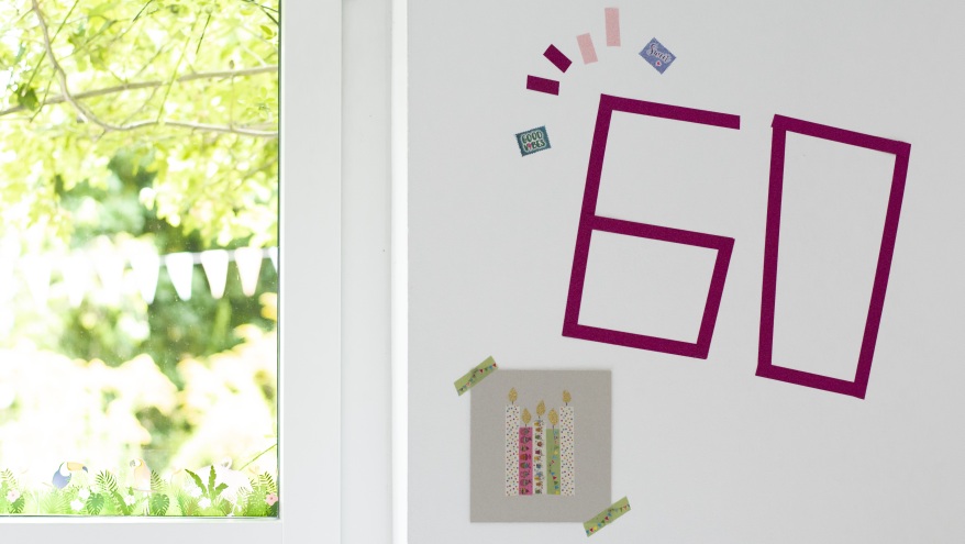 Eine weiße Wand wurde mit Washitape und einer Geburtstagskarte mit Kerzen darauf beklebt. Mit Hilfe von Washitape wurde eine 60 auf die Wand geklebt. Die Karte zeigt vier brennende Kerzen, die auch aus Washitape beklebt wurden. Im rechten Bildrand sieht man ein Fenster, das unscharf den Blick auf einen Garten freigibt. 