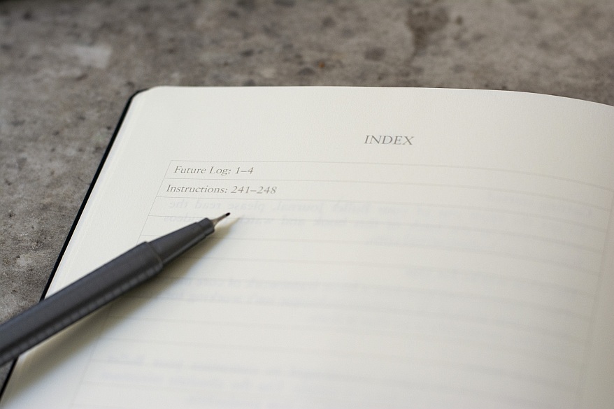 Nahaufnahme eines aufgeschlagenen Notizbuiches. Die Seite zeigt ein Index, auf der Seite liegt ein offener Fineliner.