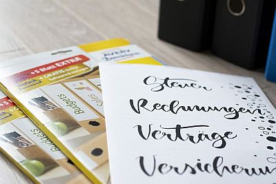 Auf einem Holztisch liegen zwei Verpackungen von Ordenerrückenetiketten von Avery Zweckform. Daraf liegt ein Bogen mit fertig gedruckten Etiketten. In der rechten, oberen Bildecke sieht man verschiedene Ordner stehen.