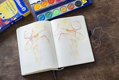 Auf einem Holztisch liegt ein aufgeschlagenes Notizbuch. Auf beiden Seiten wurden mit einem Faden gelbe und orange Mustrer gezogen. Über dem Notizbuch liegt auf dem Tisch ein augeklappter K12 Farbkasten der Marke Pelikan und die Verpackung des Fabrkastens ist zu sehen.