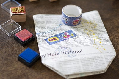 Auf einem dunklen Holztisch liegt einer zu einer Tüte gefalteten Zeitung. Diese wurde mit Maskingtape und Stempeln verziert. Daneben liegen geöffnete Stempelkissen und ein benutzter Stempel.