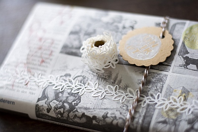 Auf einem dunklen Untergrund liegt ein Geschenk, dass mit Zeitungspapier verpackt wurde. Es wurde zusätzlich mit einem Washitape verziert, dass die Form einer Bordüre aus Blättern hat. Um das Geschenk ist Bäckergarn in gold-weiß gebunden. Daran hängt ein Geschenketikett aus Kraftpapier, welches mit einem weißen Stempel verziert wurde.