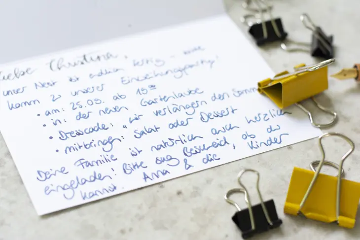 Eine aufgeklappte, beschriebene Karte mit einer Einladung zu einer Party sowie mehrere Foldback-Klammern liegen auf einem Betontisch.