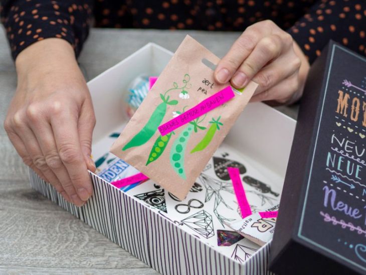 Auf einem hellen Tisch liegt eine geöffnete Box. Der Deckel ist schwarz und wurde mit Metallicmarkern belettert. Zwei Hände sind zu sehen. Eine Hand hält den Rand der Box fest, die andere hält ein Päckchen mit Erbsensamen. In der Kiste sind noch weitere Kleinigkeiten zu sehen.