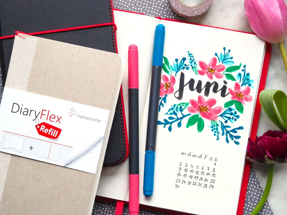 Ein geschlossenes Bullet Journal und eine mit Blumen beletterte Seite eines Bullet Journals auf einem Tisch.