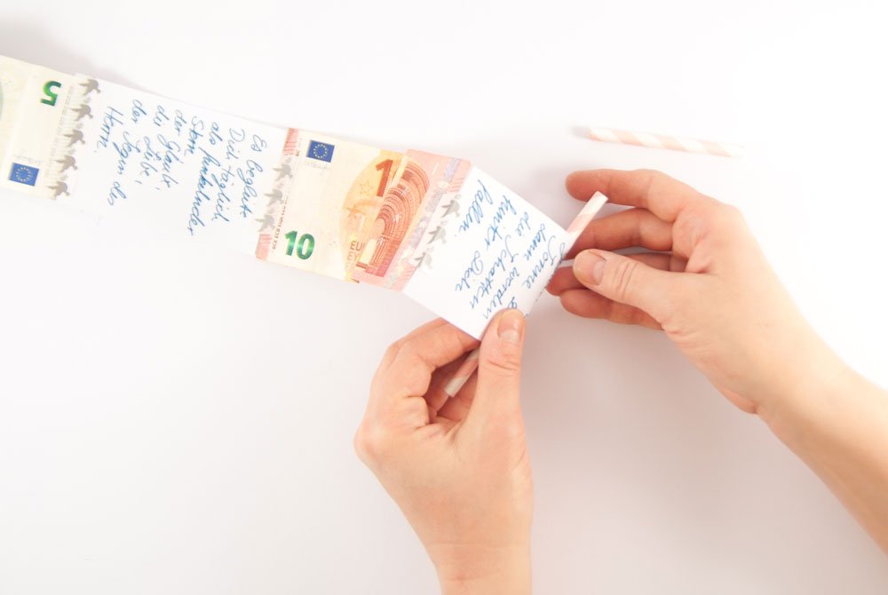Zwei Hände wickeln eine lange Reihe aus Geldscheinen und beschrifteten Papieren mit Segenswünschen darauf auf einen Papierstrohhalm. Im Hintergrund liegt ein bgeschnittenes Stück des Papierstrohhalms.