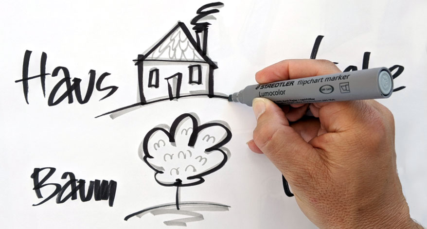 Jemand malt mit einem Staedtler Lumocolor Flipchart Marker Skizzen von Haus und Baum auf ein Blatt.