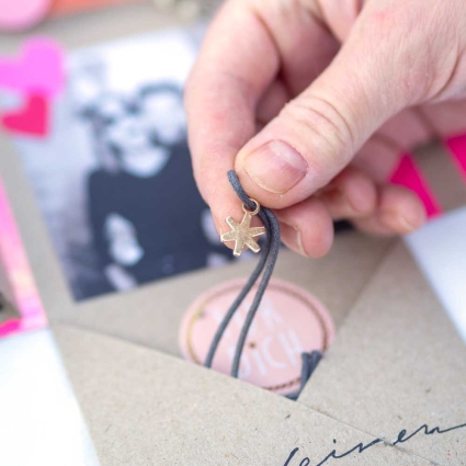 Eine Hand hält eine Kette, die in einem Umschlag steckt. Im Hintergrund ist unscharf ein Foto mit zwei Personen erkennbar.