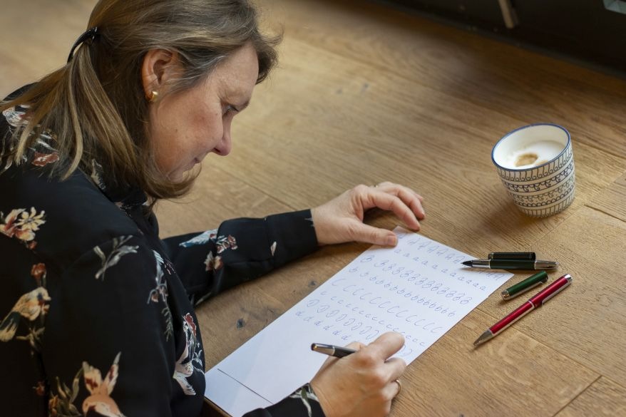 Frau sitzt am Schreibtisch und übt Schönschreiben. Sie schreibt auf ein Übungsblatt ein Alphabet, neben ihr liegen Stifte, Schreibgeräte und eine Kaffeetasse.