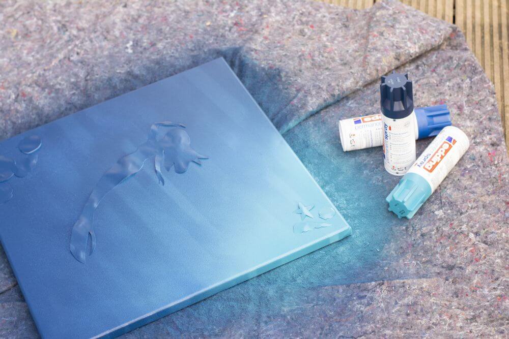 Eine Leinwand mit Schablonen in Form einer Meerjungfrau und Muscheln liegt auf einer Malerdecke. Die Leinwand wurde mich Sprühfarbe besprüht, die Sprühdosen von Edding liegen daneben.
