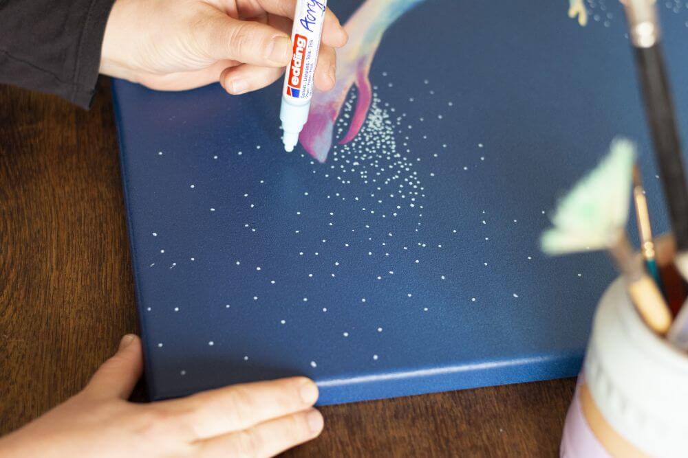 Eine Hand punktet mit einem Acrylmarker von Edding hellblaue Punkte auf eine Leinwand in dunkelblauer Farbe.