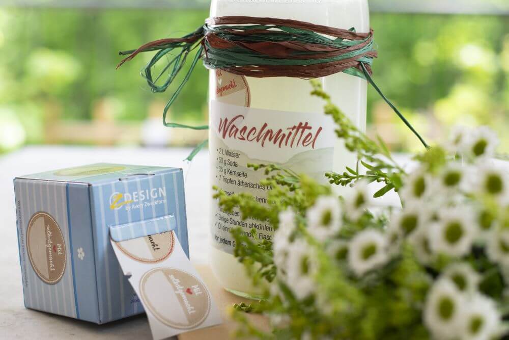 Detailaufnahme von einer Glasflasche mit selbstgemachtem DIY-Waschmittel. Daneben stehen Sticker von der Rolle von Avery Zweckform, im Vordergrund liegt ein Blumenstrauß.