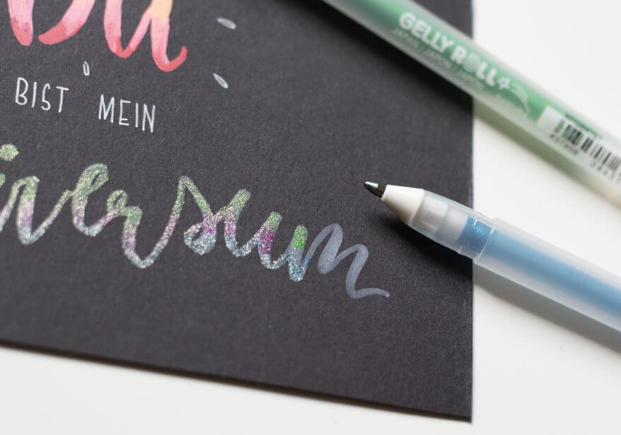 Nahaufnahme eines Handletterings auf schwarzem Papier: Mit glitzernden Gelstiften "Gelly Rolls Stardust" von Sakura verschwimmen die Farben in einem Blending im Wort "Universum".