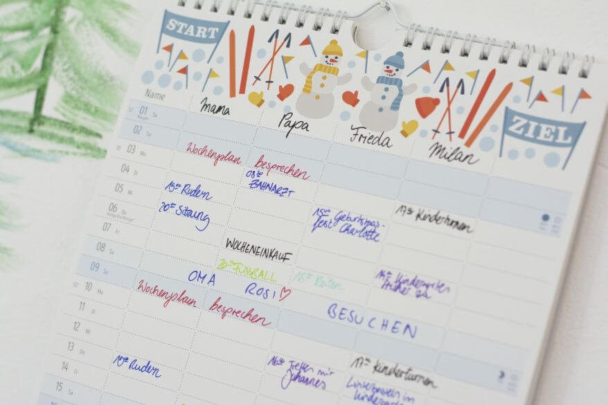 Ein beschriebener Familienplaner von Heye hängt, mit verschiedenen Terminen und Verabredungen beschriftet, an der Wand.
