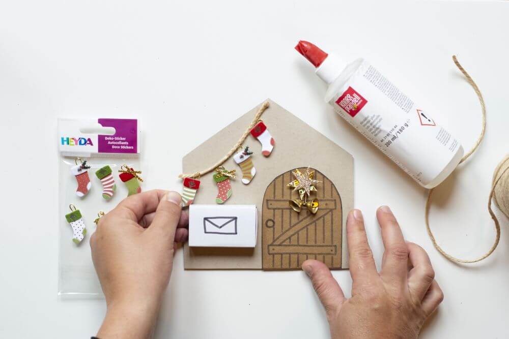 Wichteltür zu Weihnachten basteln, Bastelanleitung Schritt 6: Mit Hilfe von Dekostickern in Sockenform der Marke Heyda eine Leine mit Weihnachtssocken auf das Wichtelhaus kleben.