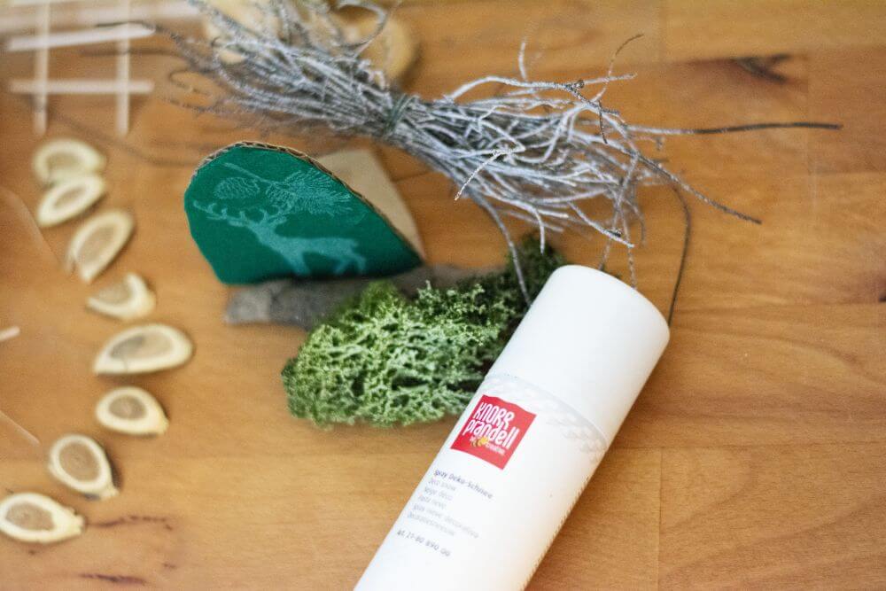 Auf Parkettboden liegend sieht man verschiedene Dekomaterialien, mit denen eine Wichteltür zur Weihnachtszeit ergänzt wird: kleine Baumscheiben, ein gestempelter Hirsch, sowie Moos und Äste, die mit Dekoschnee von Knorr Prandell besprüht wurden.