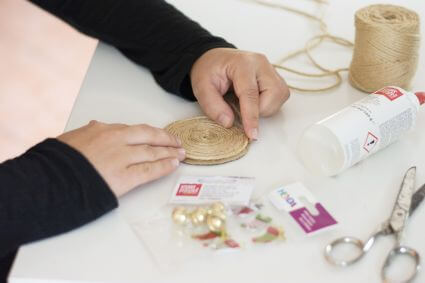 Blick auf einen weißen Schreibtisch, wo zwei Hände mit Hilfe von Jutegarn und Flüssigkleber von Knorr Prandell einen kleinen Teppich für eine selbstgemachte Wichteltür basteln.