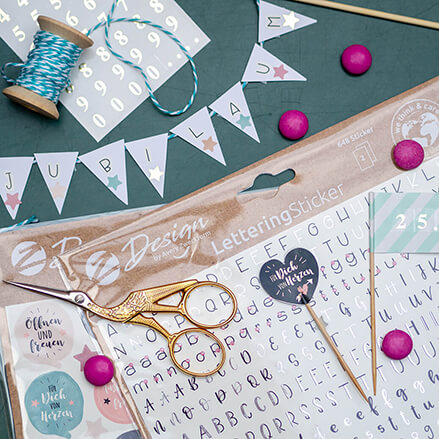 Zahlreiche (Lettering-) Sticker liegen auf einem Tisch. Außerdem sieht man eine Schere, Perlen, Farn und eine Girlande, auf der "Jubiläum" steht.