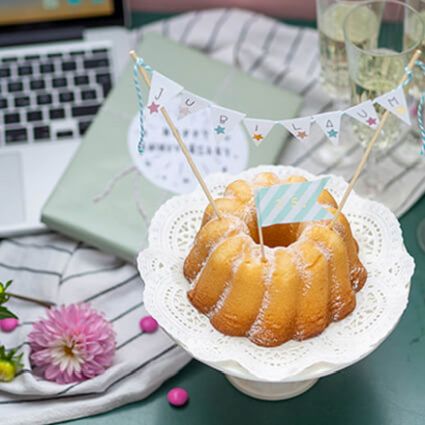 Ein Gugelhupf mit „Jubiläum“-Girlande auf einer Tortenplatte, daneben stehen ein Laptop, ein Geschenk und Sektgläserr.