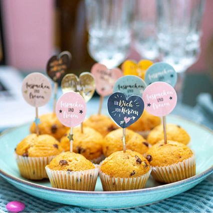 Ein Teller mit Schoko-Muffins, dekoriert mit Spruch-Steckern asu Stickern von Avery Zweckform, im Hintergrund  stehen Sektgläser.