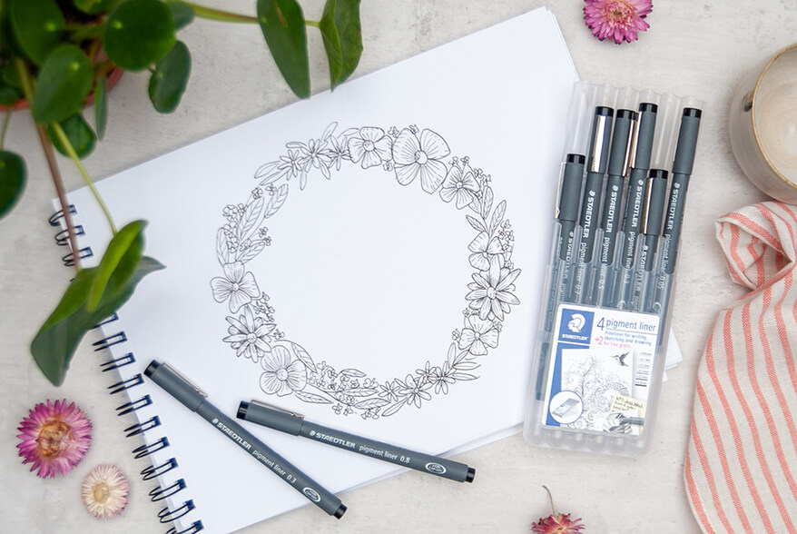 Auf einem hellen Untergrund liegt ein Zeichenblock. Darauf ist mit schwarzem Pigment Liner ein Blumenkranz gezeichnet. Auf und neben der Zeichnung liegen weitere Pigment Liner von Staedtler, teilweise in ihrer Verpackung. Eine Grünpflanze am linken oberen Rand des Bildes, sowie rosafarbene Trockenblumen lockern das Bild auf. Foto: Johanna Rundel