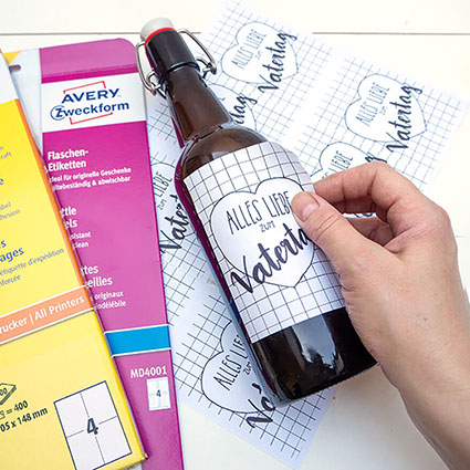 Jemand klebt ein selbstgestaltetes Etikett mit einem Glückwunsch zum Vatertag auf eine Bierflasche. Daneben liegen Verpackungen mit verschiedenen Blanko-Etikettbögen von Avery Zweckform. Foto: Johanna Rundel